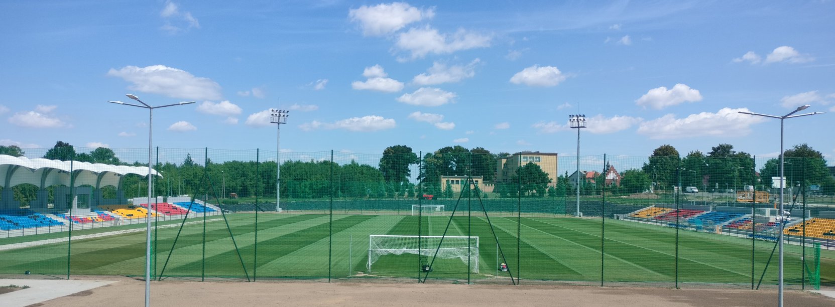 Stadio, widok z góry od strony bramki. Po bokach kolorowe trybuny, w tle zieleń drzew