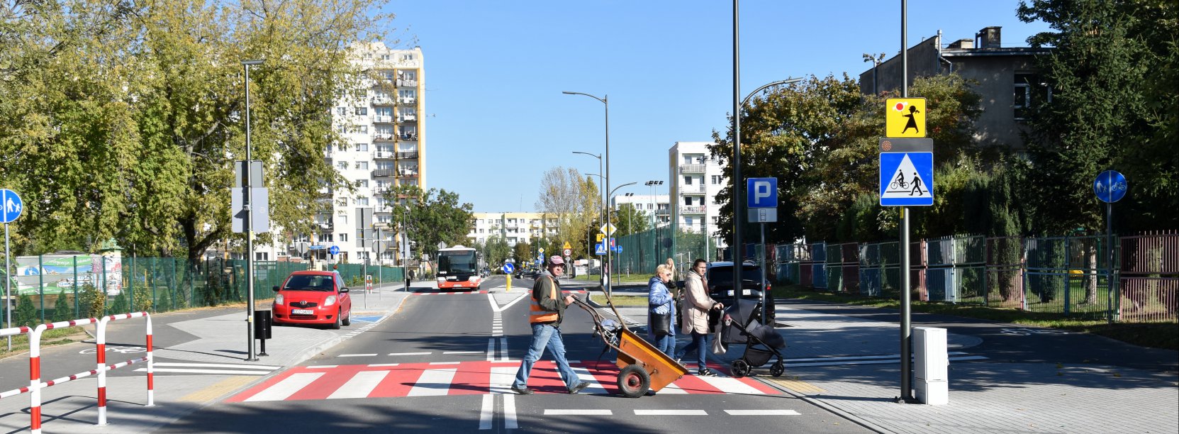 Trzy osoby przechodzące przez biało-czerwone przejście dla pieszych, w tle wieżowiec