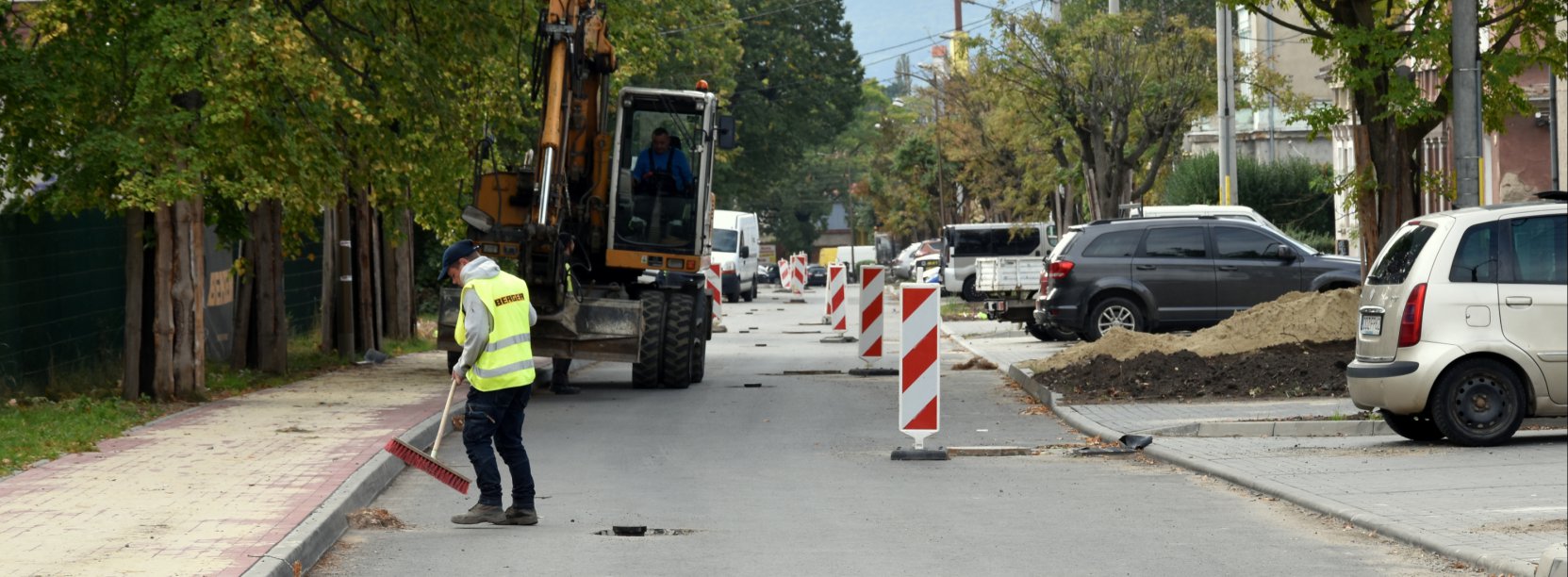 Prace przy budowie drogi, człowiek w jaskrawej kamizelce zamiata drogę, za nim koparka