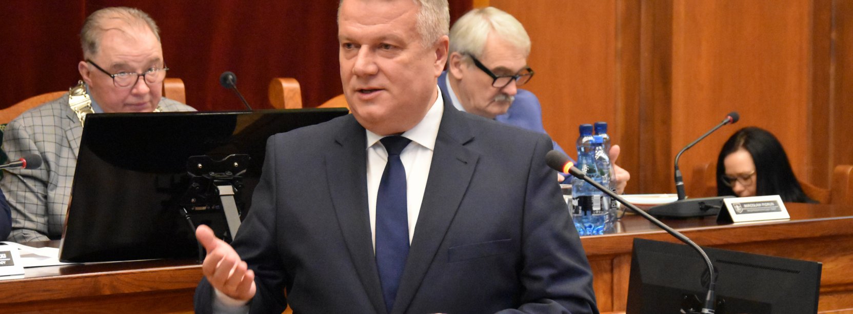 Burmistrz przy mównicy, za nim dwie osoby przy stole prezydialnym 