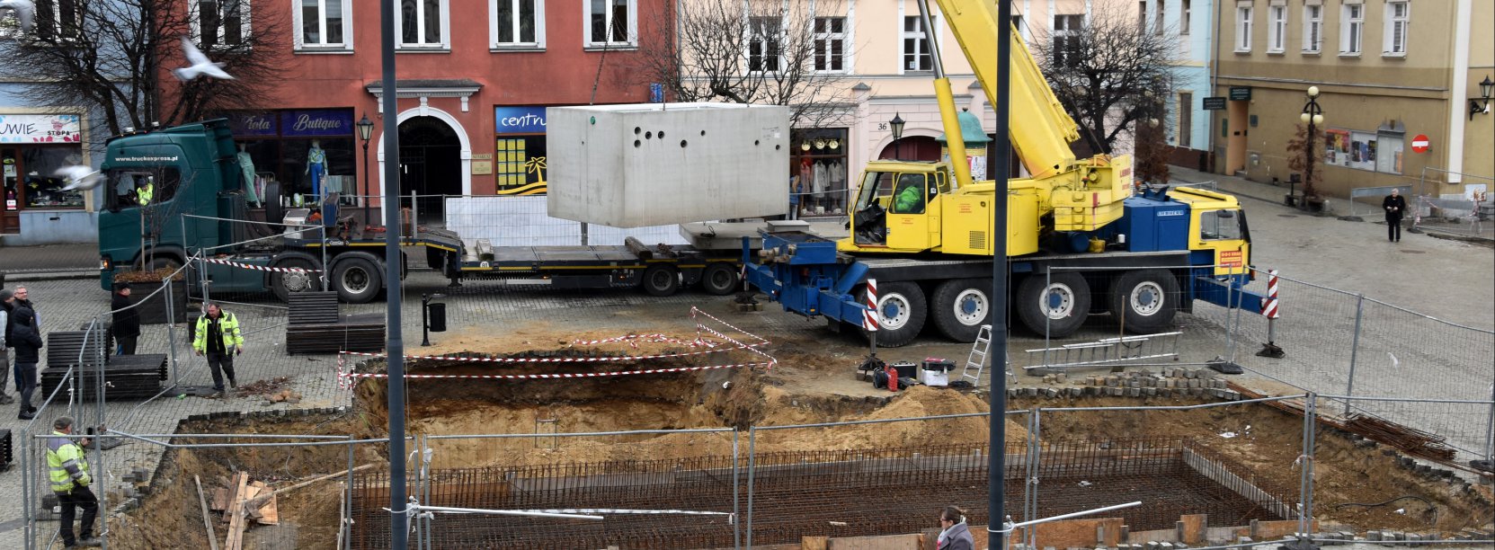 Dźwig unoszący dużą betonową konstrukcję, w tle kamienice