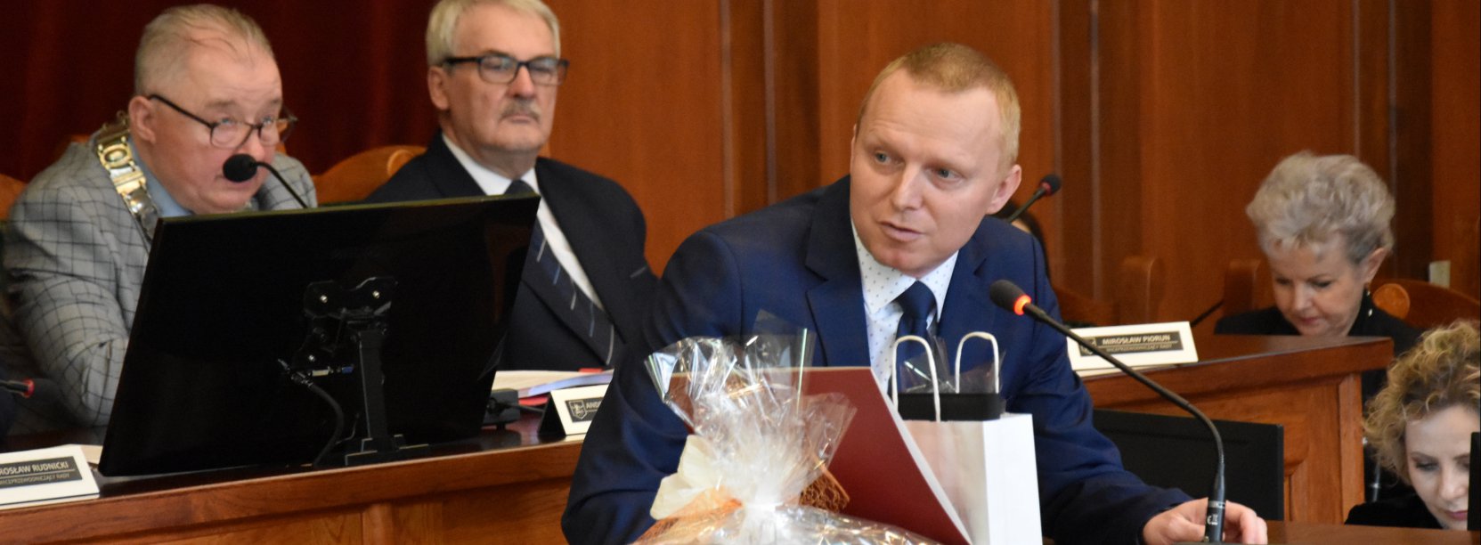 Mężczyzna stojący przy mównicy, za nim stół z dużym monitorem i dwiema osobami