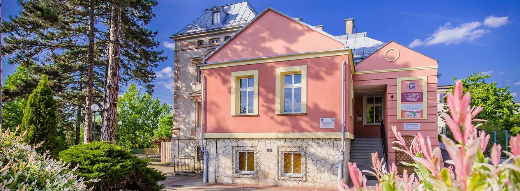 Kamienica z różową elewacją, dookoła zieleń i kwiaty, błękitne niebo 
