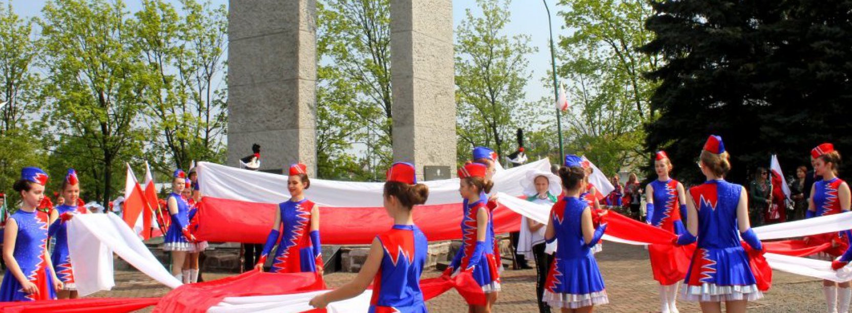 Kilkanaście dziewczyn w niebieskich strojach trzyma szarfy w barwach flagi Polski, za nimi pomnik z metalowym godłem