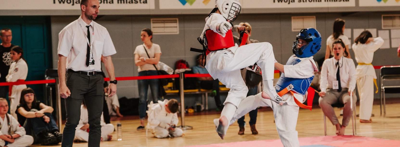 Karateka z wyskoku atakuje przeciwnika, obok sędzia 