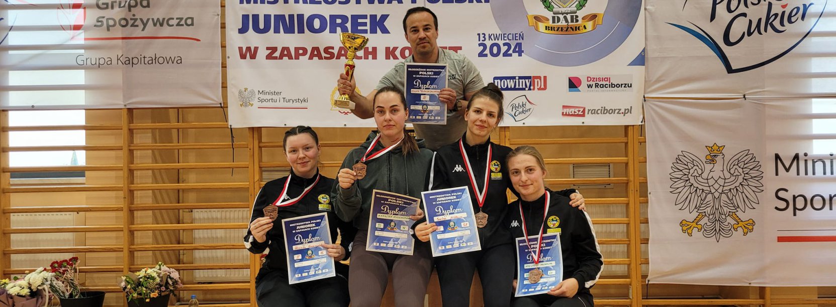 Cztery zawodniczki z dyplomami siedzące na podium, z nimi trener z pucharem w ręku