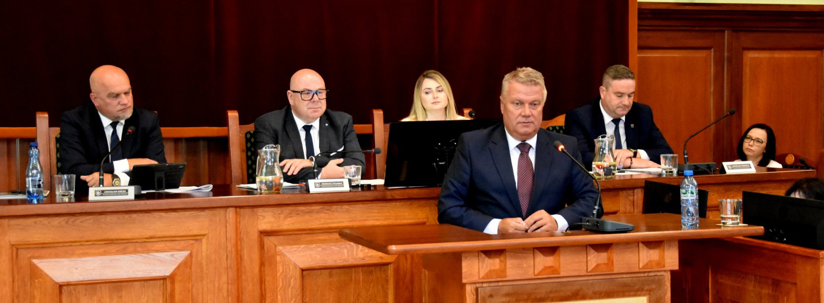 Burmistrz przy mównicy, za nim cztery osoby siedzące przy stole prezydialnym