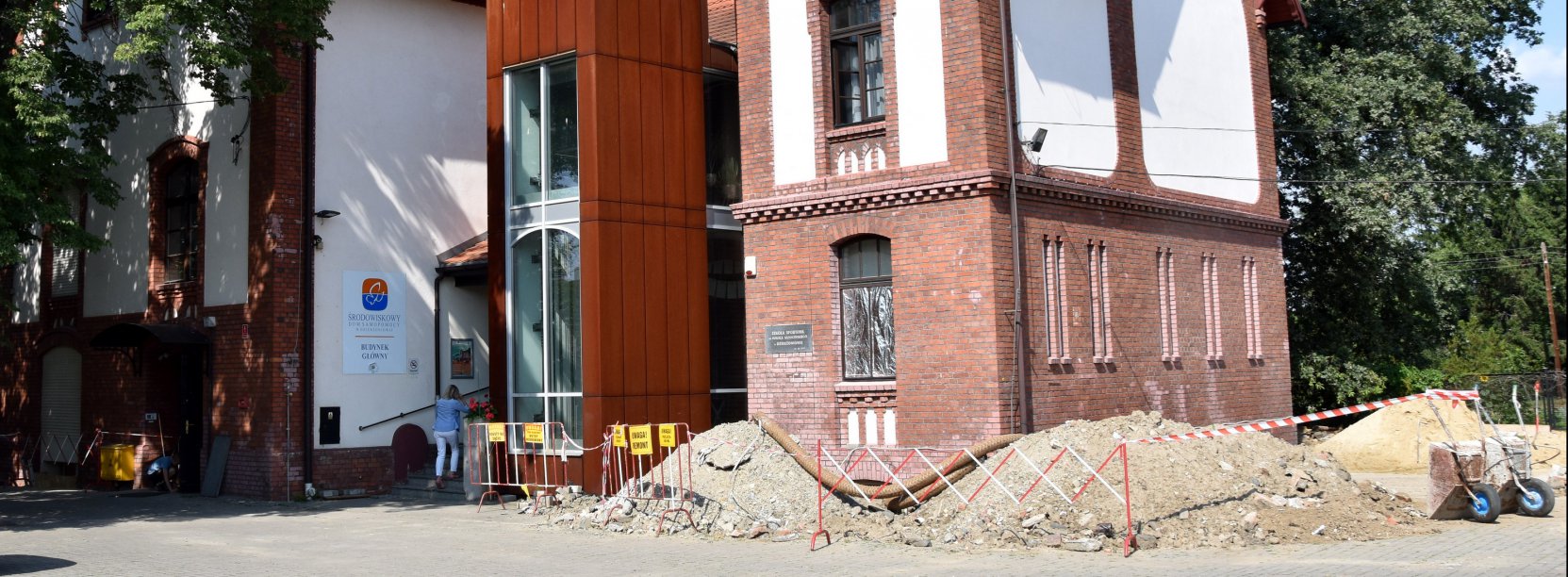 Kamienica z cegiegieł i białej elewacji, przed nią hałdy ziemii i narędzia budowlane