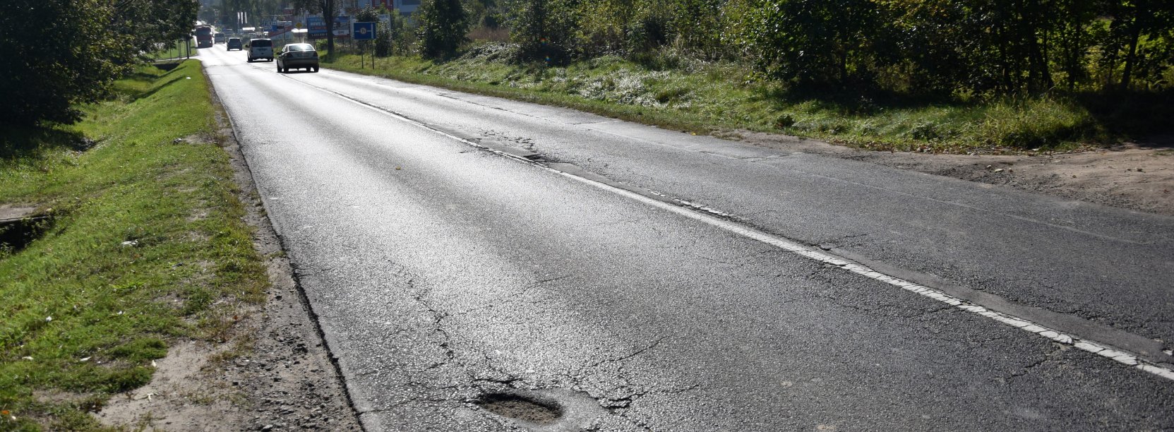 Droga asfaltowa prowadząca do miasta, w dali samochody, po bkach drzewa i krzewy