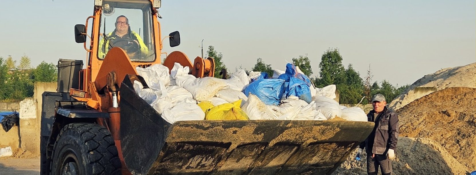 Worki z piaskiem na "łyżce" dużej koparki, obok hałdy piasku