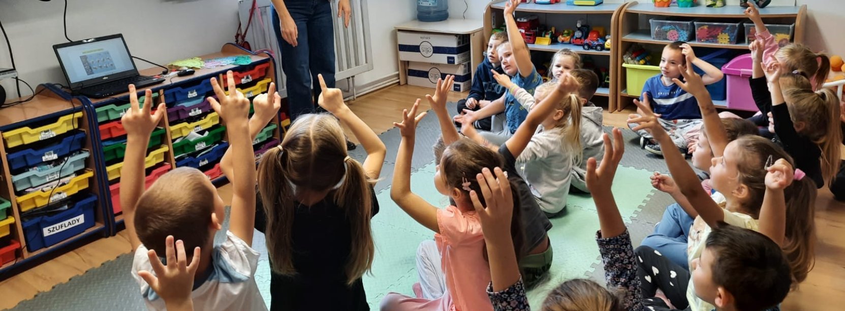 Przedszkolaki siedzący w grupie zgłaszają się do odpowiedzi, przed nimi młoda kobieta