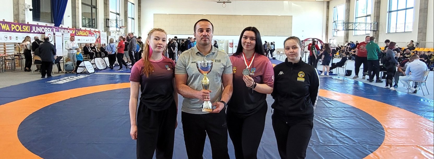 Trener z trzema zawodniczkami, za nimi mata do zapasów i tłum sportowców