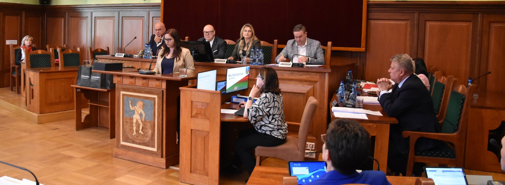 Sala obrad, za stopłem prezydialnym siedzą cztery sosoby, przed stołem mównica