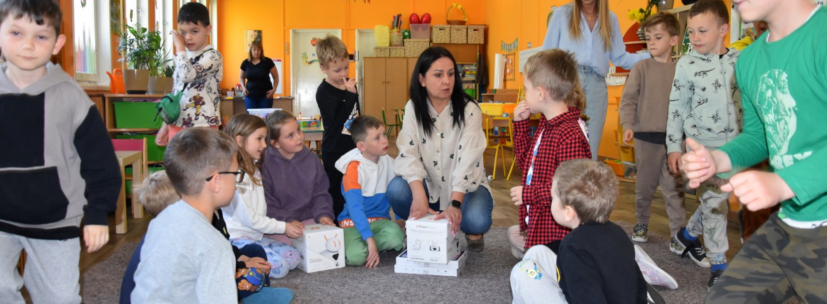 Grupa przedszkolaków w dużej sali, siedzących na podłodze i rozpakowujących z pudełek roboty i tablety