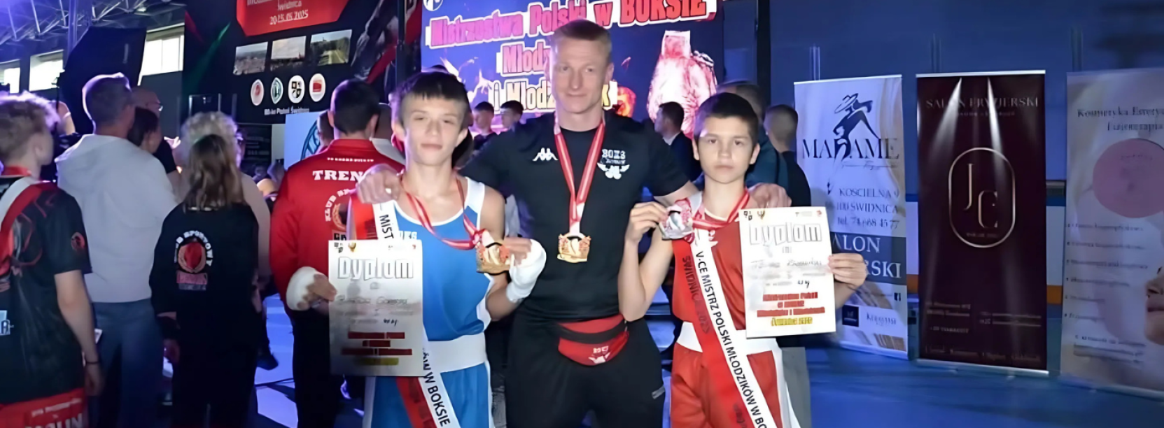 Dwóch młodych zawodników w sportowych strojach, z dyplomami i medalami, pomiędzy nimi trener
