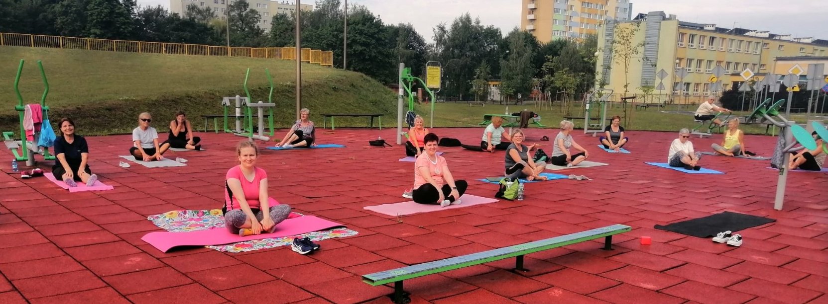 Kilkanaście osób siedzących na matach do ćwiczeń na terenie zewnętrzej siłowni, za nimi zieleń i bloki osiedlowe