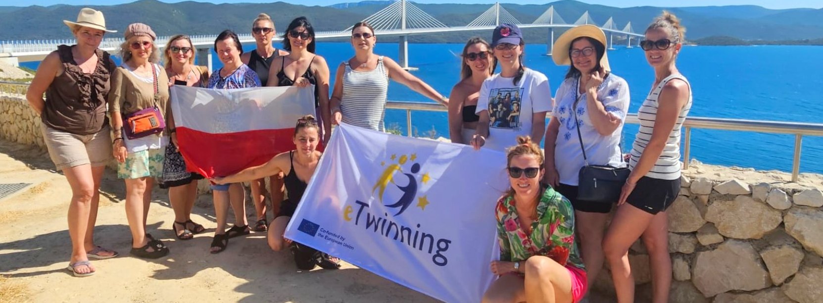 Nauczycielki z flagami Polski i projektu Twinning na tle morza, w dalekim planie duży most i góry