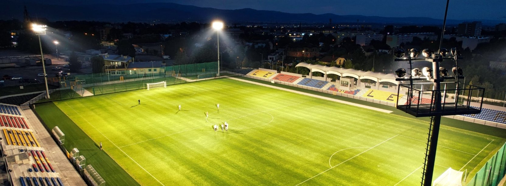 Stadion piłkarski nocą, oświetlony, z trybunami po bokach, na muraiwe grupka piłkarzy