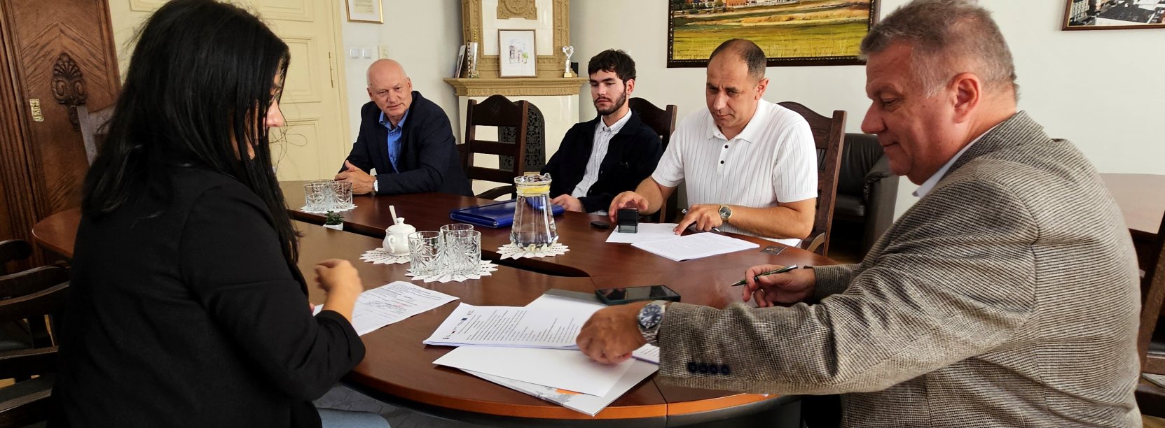 Zdjęcie przedstawiające siedzących przy stole czterech mężczyzn i kobietę. Dwóch z mężczyzn podpisuje dokumenty
