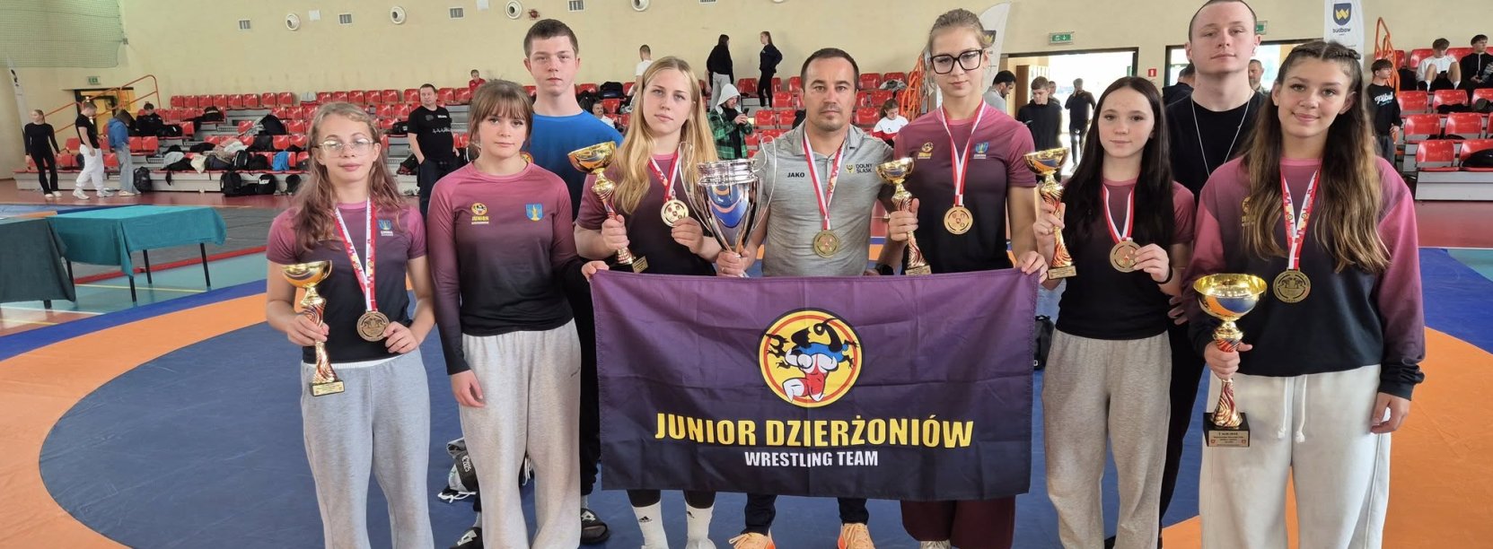 grupa 5 zapaśniczek z medalami i pucharami z trenerami i flagą klubu sportowego Junior Dzierżoniów