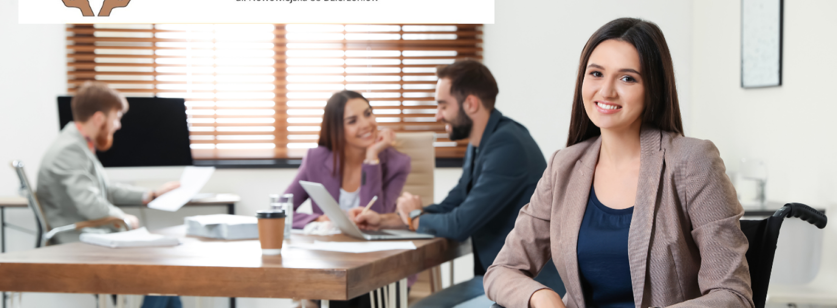 Biuro, osoby rozmawiające przy stole, na pierwszym planie uśmiechnięta, siedząca na wózku niepełnosprawna kobieta