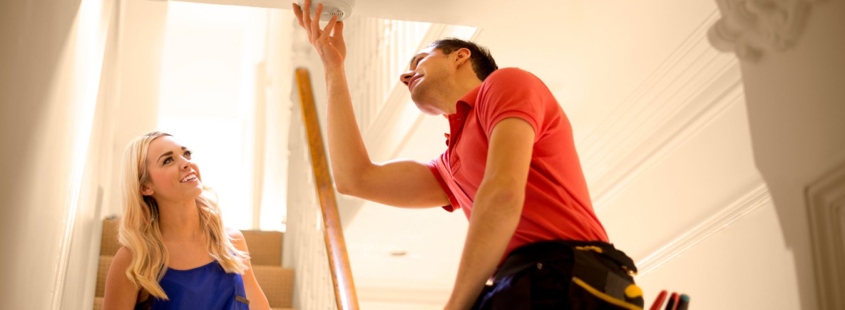 Wneętrze domu, mężczyzna montujący czujkę dymu. Przygląda się temu stojąca na schodach kobieta