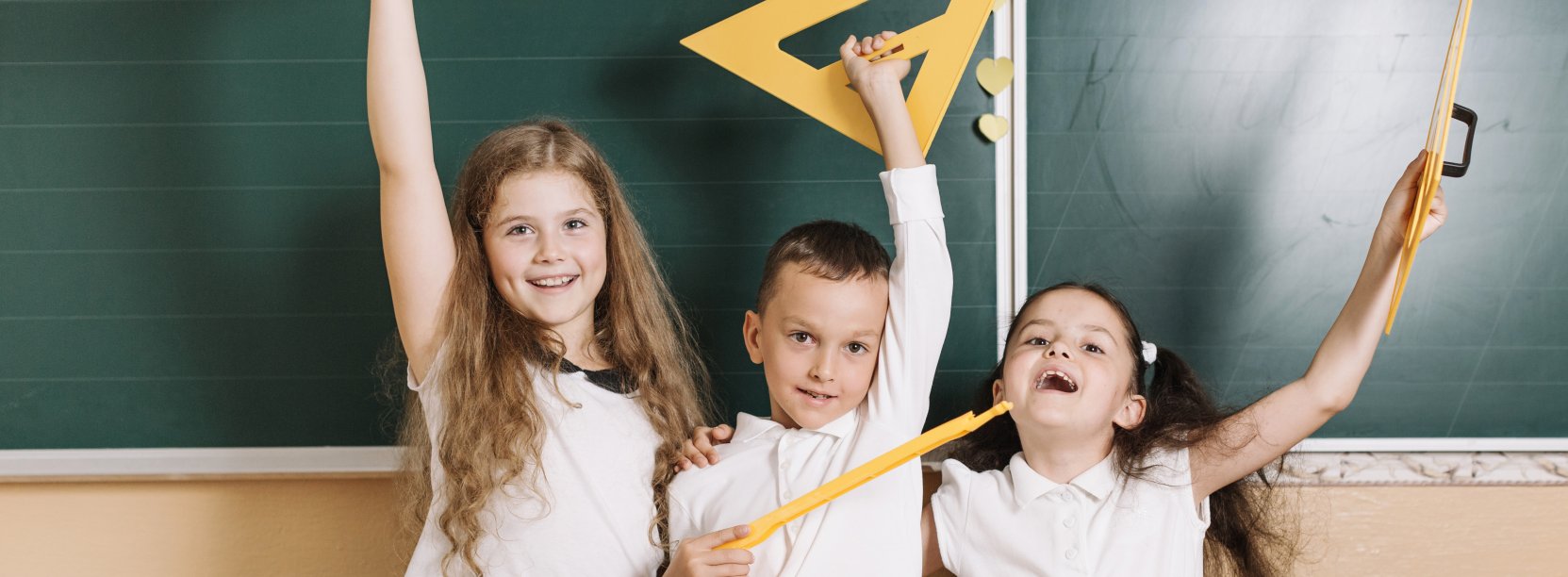 troje dzieci z przyrządami do geometrii stoi przed tablicą w klasie