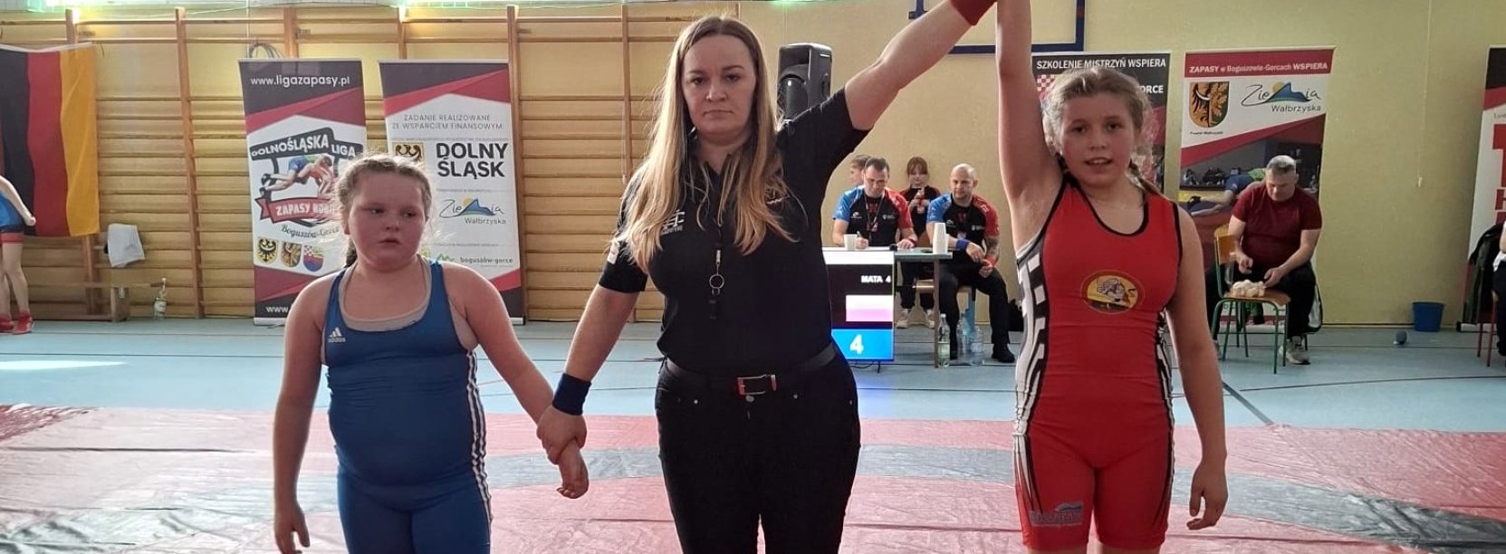 Dwie zawodniczki i sędzina na macie podczas ogłaszania wyniku