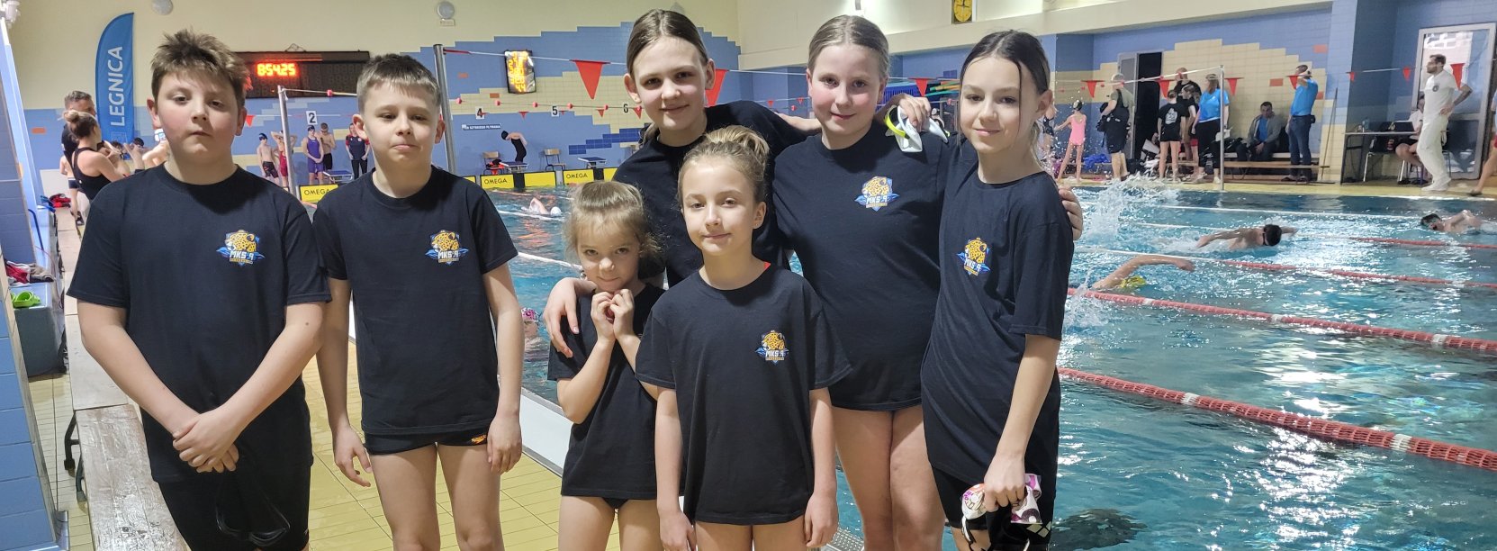 Siedmioro młodych pływaków w jednakowych granatowych koszulkach przy krawędzi basenu