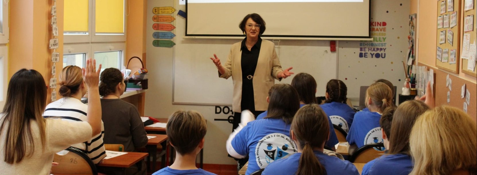 Uczniowie w ławkach przy tablicy kobieta prowadząca zajęcia