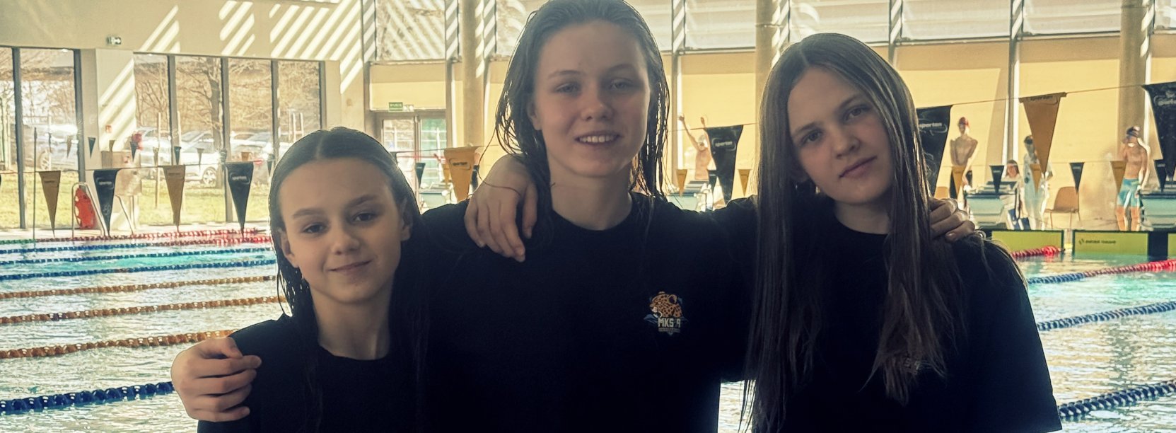 Trzy młode zawodniczki w czarnych koszulkach na tle basenu