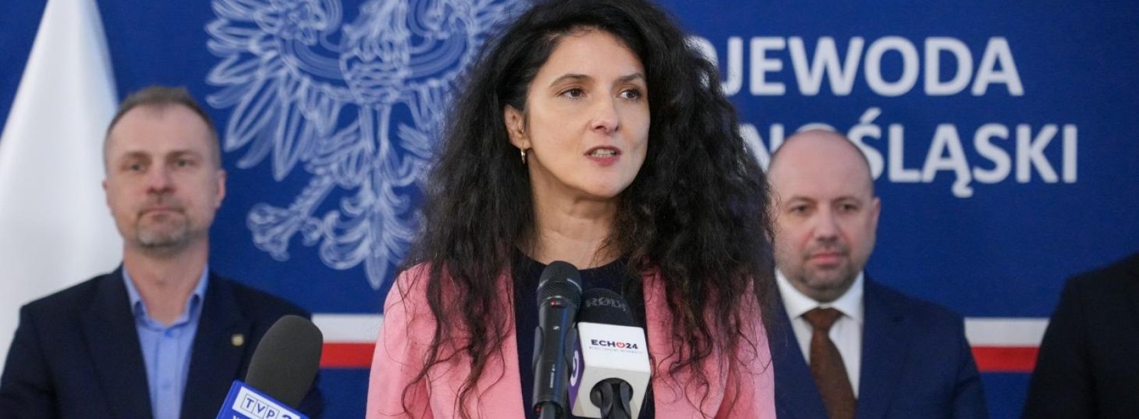 Wojewoda Dolnośląska przy mikrofonie, za nią dwóch mężczyzn na niebieskim tle