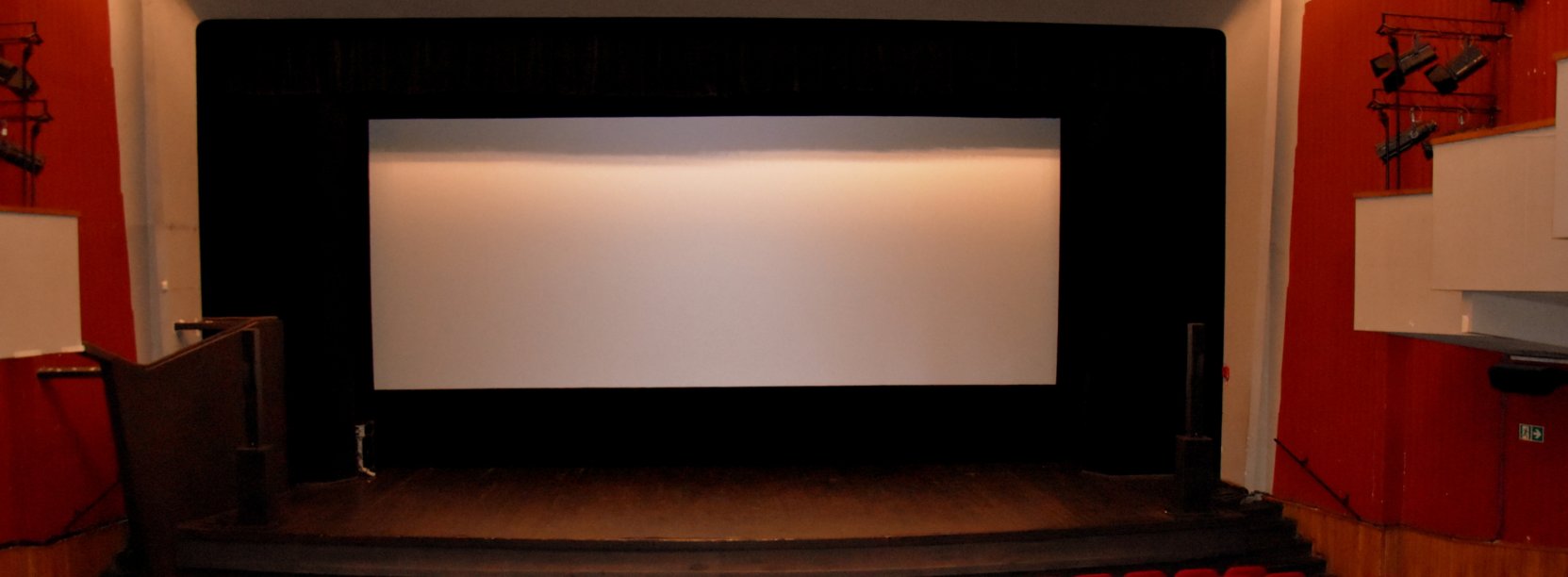 Biały ekran kinowy, przed ekranem scena i czerwone krzesełka, po bokach czerwone ściany