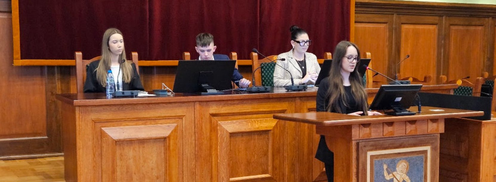 Młodzieżowi radni, dziewczyna stojąca przy mównicy, chłopak i dwie dziewczyny siedzący za stołem prezydialnym