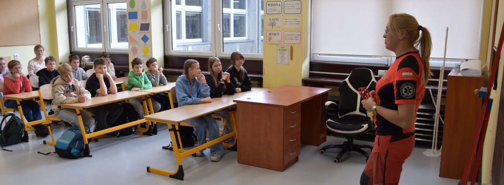 Pracownik oddziału ratunkowego w pomarańczowo-czarnym stroju stoi w klasie przed uczniami siedzącymi w ławkach