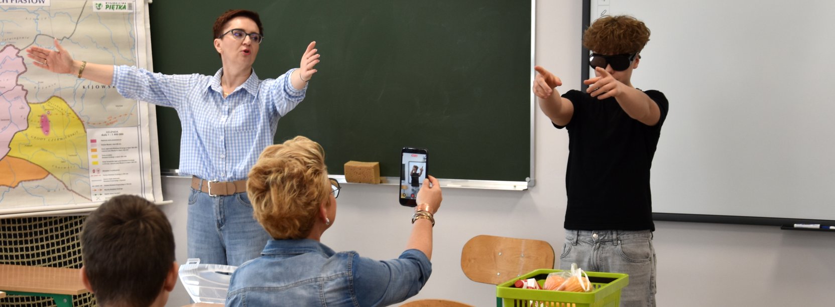 Prowadząca zajęcia przy szkolnej tablicy z rozłożonymi rękoma, obok uczeń w ciemnych okularach wskazujący na drugi koniec sali