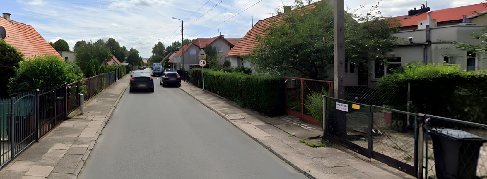 Uliczka, zaparkowane i jadące samochody, po bokach małe domki jednorodzinne