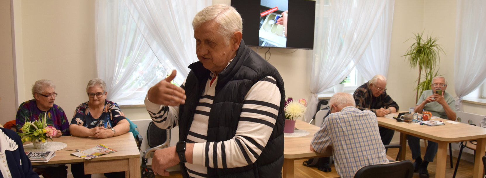 Senior z opaską na ręce, za nim przy stołach siedzi grupa seniorek i seniorów