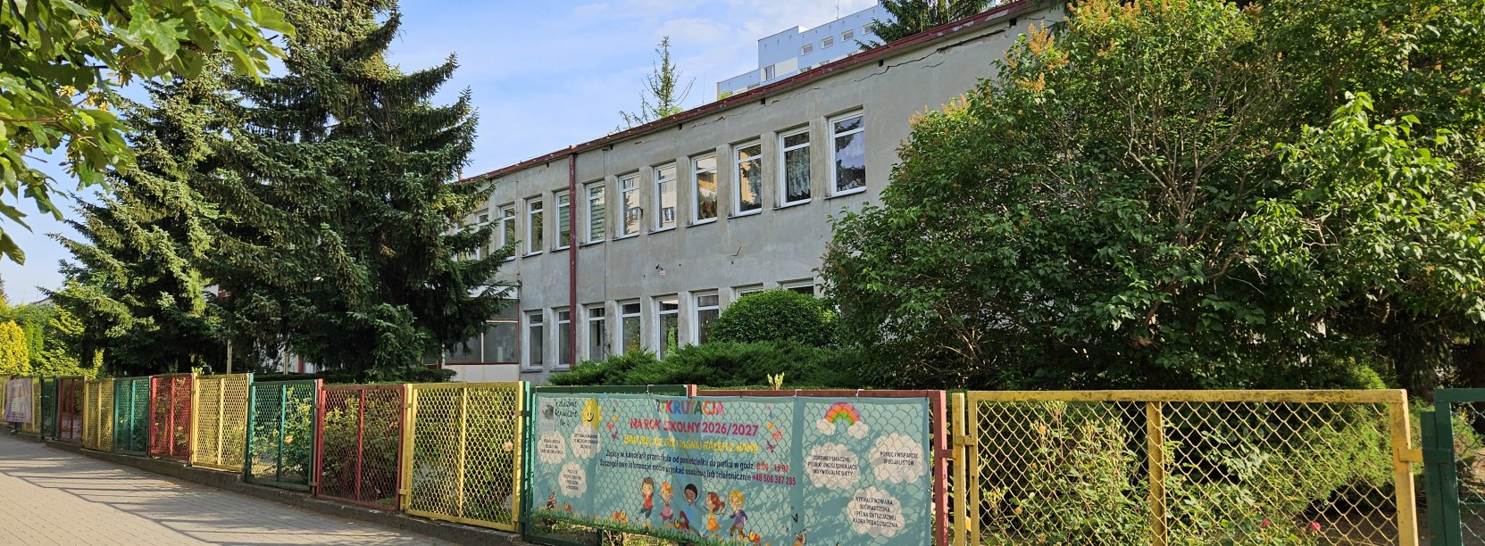 Dwupiętrowy długi budynek częściowo zasłonięty drzewami, przed budynkiem kolorowe ogrodzenie