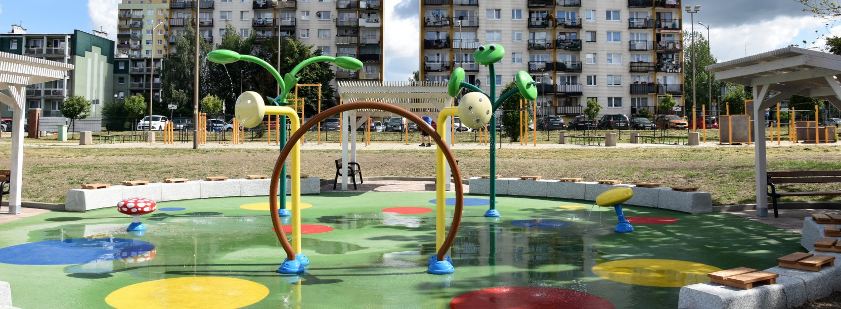 Wodny plac zabaw z zielona nawierzchnią i z urządzeniami tryskającymi wodę, w tle dwa wieżowce