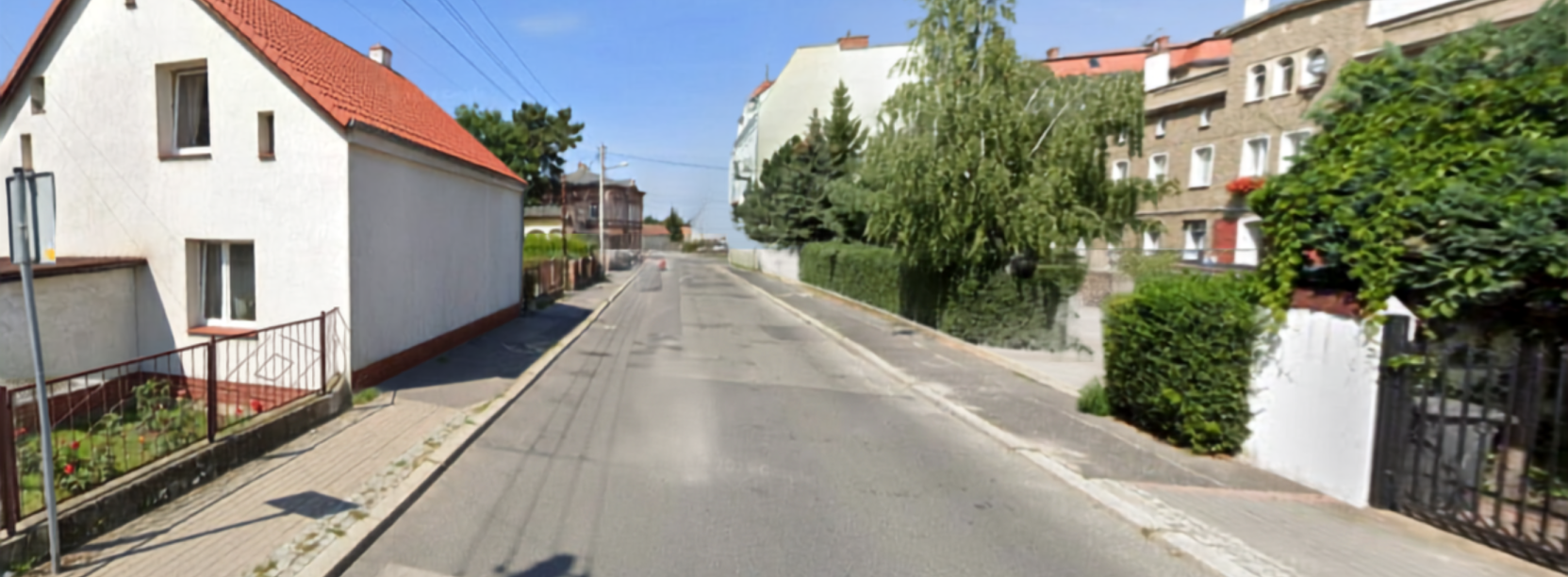 Droga asfaltowa, po lewej biały domek jednorodzinny, po praweej drzewa i domy jednorodzinne