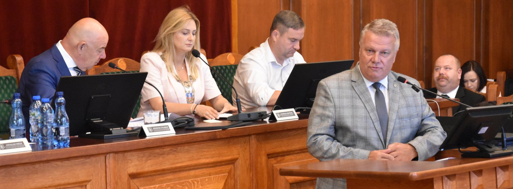 Burmistrz Dzierżoniowa przy mównicy, za nim przy stole prezydialnym przewodnicząca i dwóch wiceprzewodniczących rady