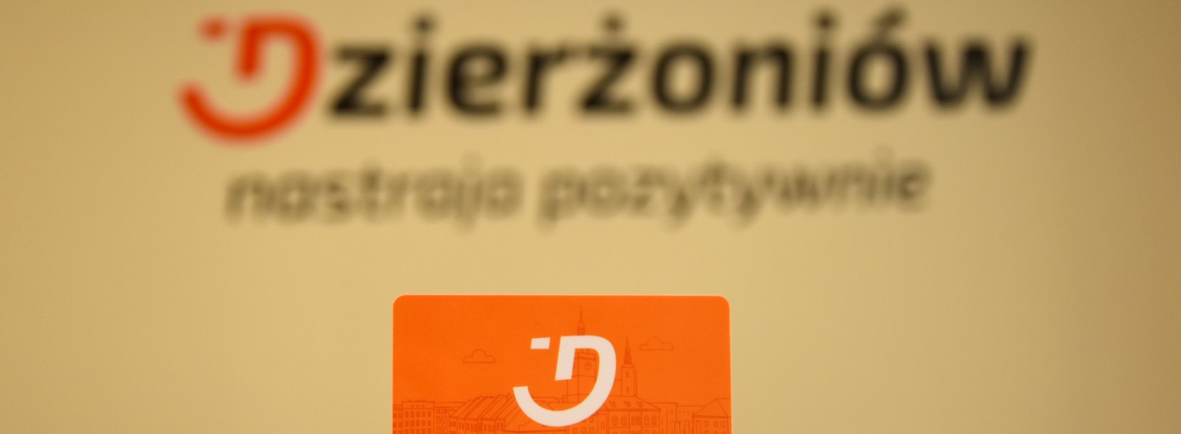 Dłoń trzymająca pomarańczową kartę mieszkańca na tle logotypu miasta