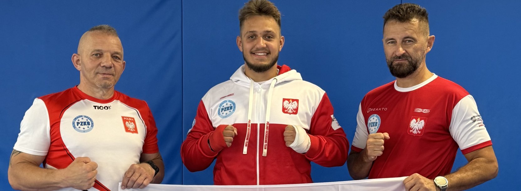 Maksymilian Pelej w biało-czerwonym dresie. Po bokach trenerzy trzymający flagę z logotypem Dzierżoniowa