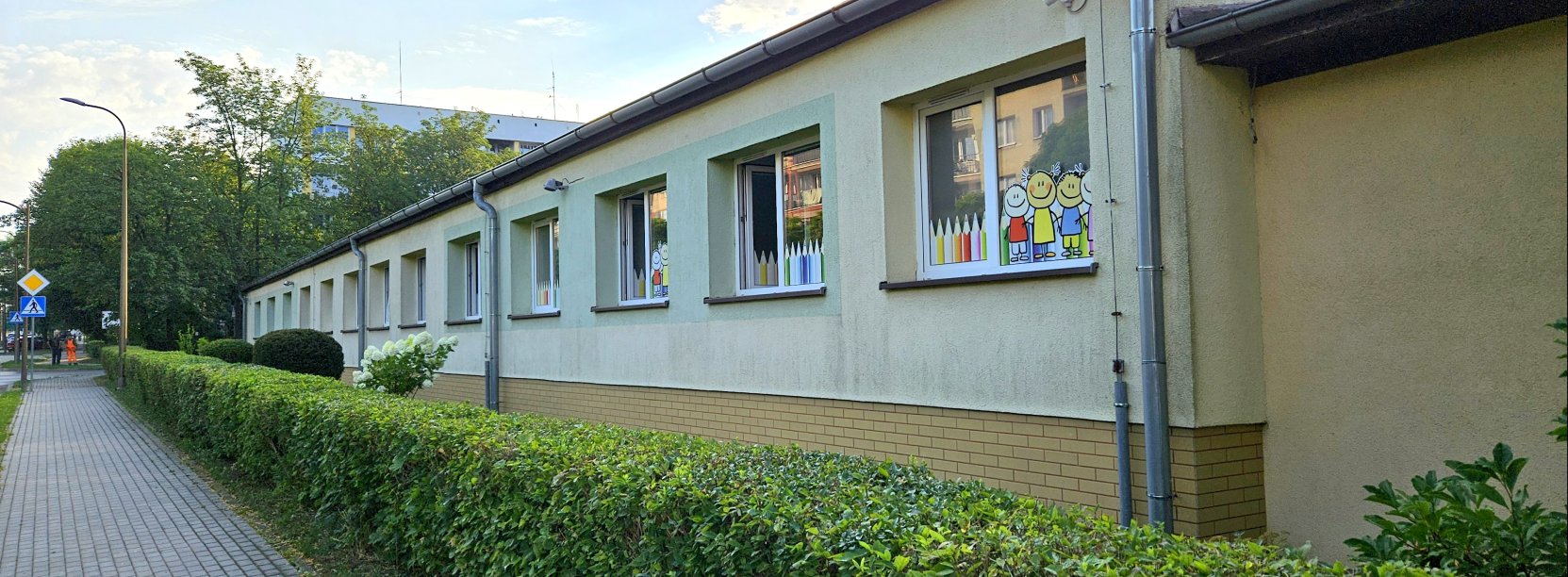 Długi parterowy budynek, przed nim żywopłot i chodnik 