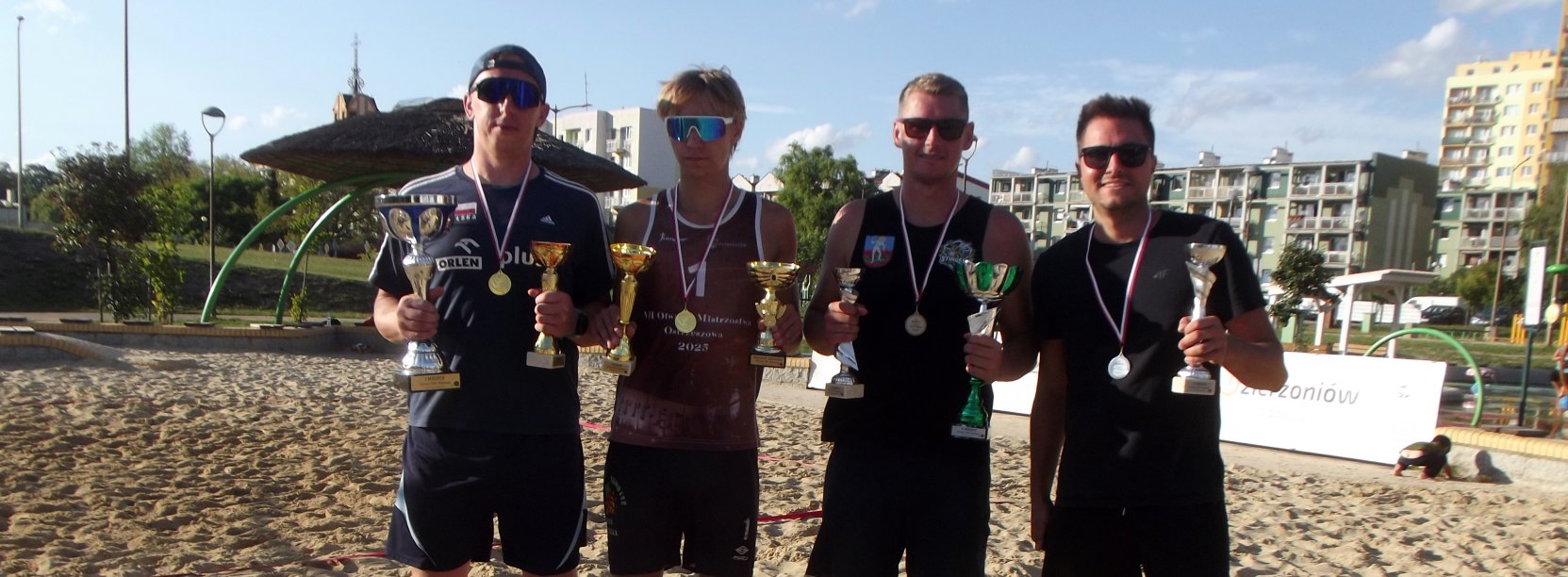 Czterech zawodników, każdy z pucharem i medalem, stojących na boisku do siatkówki plażowej, za nimi bloki osiedlowe