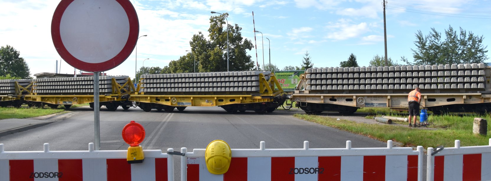 Przejazd kolejowy zagrodzony barierkami, na torach skład wiozący podkłady kolejowe