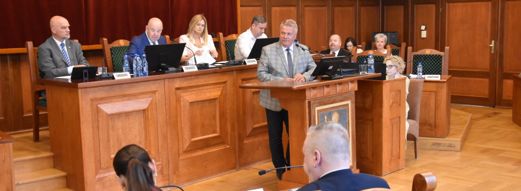 Sala obrad, przy mównicy burmistrz, za nim przewodnicząca i trójka wiceprzewodniczących przy stole prezydialnym