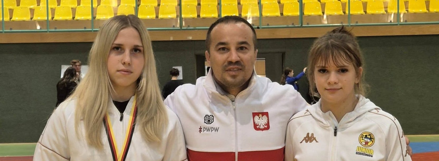 Trener w biało-czerwonej bluzie, po bokach dwie zawodniczki 