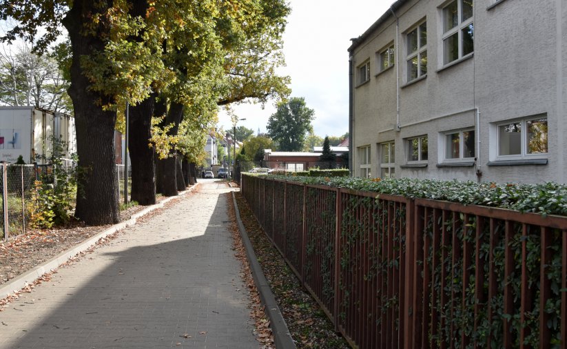 Droga z kostki betonowej, po prawej stronie ogrodzenie, żywopłot i fragment budynku